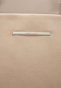 Helle beigefarbene, strukturierte Kunstlederoberfläche mit einem silbernen Metallelement, auf dem der eingravierte Markenname "ANNA FIELD" in einem minimalistischen Design zu sehen ist.