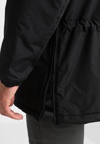 Czarna kurtka o gładkiej fakturze, z elastycznymi mankietami i bocznym zamkiem. Widoczna kieszeń na wysokości talii z marszczonym detalem.