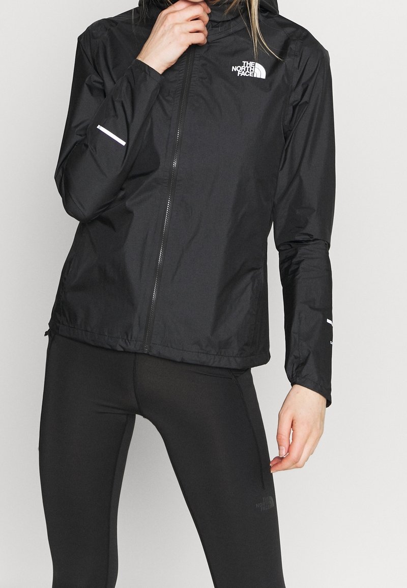 Personne portant une veste noire The North Face et des leggings noirs, ajustant le col de la veste d’une main.