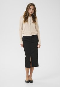 Cárdigan de punto en Beige claro con botones, combinado con una falda negra de longitud hasta la rodilla que presenta una abertura frontal. Llevado con zapatos planos negros.
