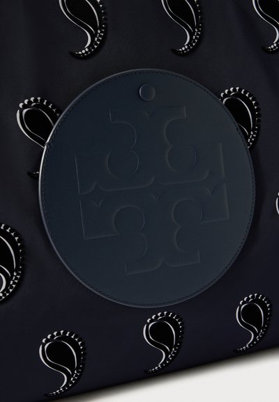 Σκούρο μπλε ύφασμα με μαύρα και λευκά σχέδια paisley που περιβάλλουν μια στρογγυλή δερμάτινη επιφάνεια με ανάγλυφο γεωμετρικό λογότυπο.