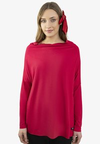 Młoda kobieta o długich brązowych włosach, ubrana w luźną czerwoną bluzkę z długim rękawem i czerwoną kokardę we włosach, stojąca na białym tle.