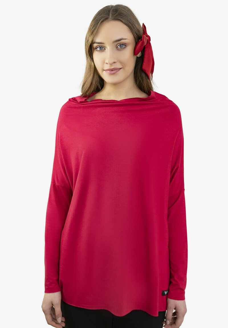 Młoda kobieta o długich brązowych włosach, ubrana w luźną czerwoną bluzkę z długim rękawem i czerwoną kokardę we włosach, stojąca na białym tle.