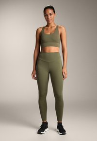 Nő áll, olívazöld sportmelltartóban és hozzá illő, magas derekú leggingsben, fekete sportcipővel, egyszínű háttér előtt.