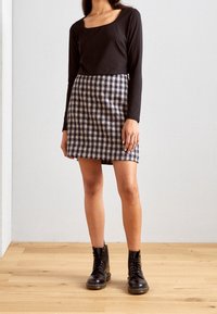 Kvinna som bär en svart långärmad topp, en rutig minikjol och svarta snörade ankelstövlar, stående på en trägolv mot en slät vägg.