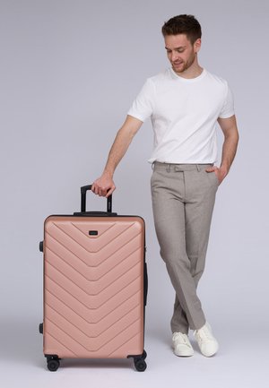 Valigia rigida color oro rosa con motivo a zigzag, dettagli neri, manico telescopico e quattro ruote per la mobilità.