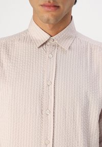 Camicia button-up di un rosa chiaro con texture, dotata di un colletto piccolo, caratterizzata da strisce verticali e cinque bottoni frontali.