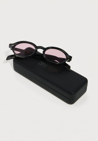 Μαύρα οβάλ γυαλιά ηλίου με ροζ φακούς που ακουμπούν σε μια υφή μαύρη ορθογώνια θήκη γυαλιών πάνω σε φωτεινή επιφάνεια.
