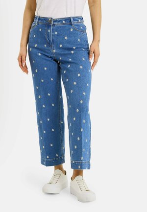 Vaqueros azules de mezclilla con pequeños patrones de margaritas blancas, usados con zapatillas blancas con plataforma y una camiseta blanca en una persona de pie.