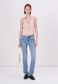 Ροζ αμάνικο τοπ με λεπτομέρεια μπροστά που δένει, ανοιχτό μπλε εφαρμοστά τζιν και μαύρες λοφάσιες. Κρατώντας μια άσπρη τσάντα. Απλό φόντο.