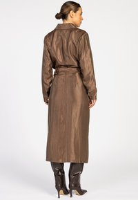 Vrouw met een haar in een knot die een glanzende lange bruine jas met riem draagt, gecombineerd met donkerbruine laarzen met hak tot aan de knie, staande tegen een witte achtergrond.