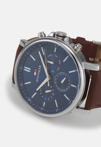 Montre chronographe avec un cadran bleu marine, des aiguilles argentées et un bracelet en cuir marron. Elle dispose d'indicateurs de jour et de date ainsi que d'accents métalliques.