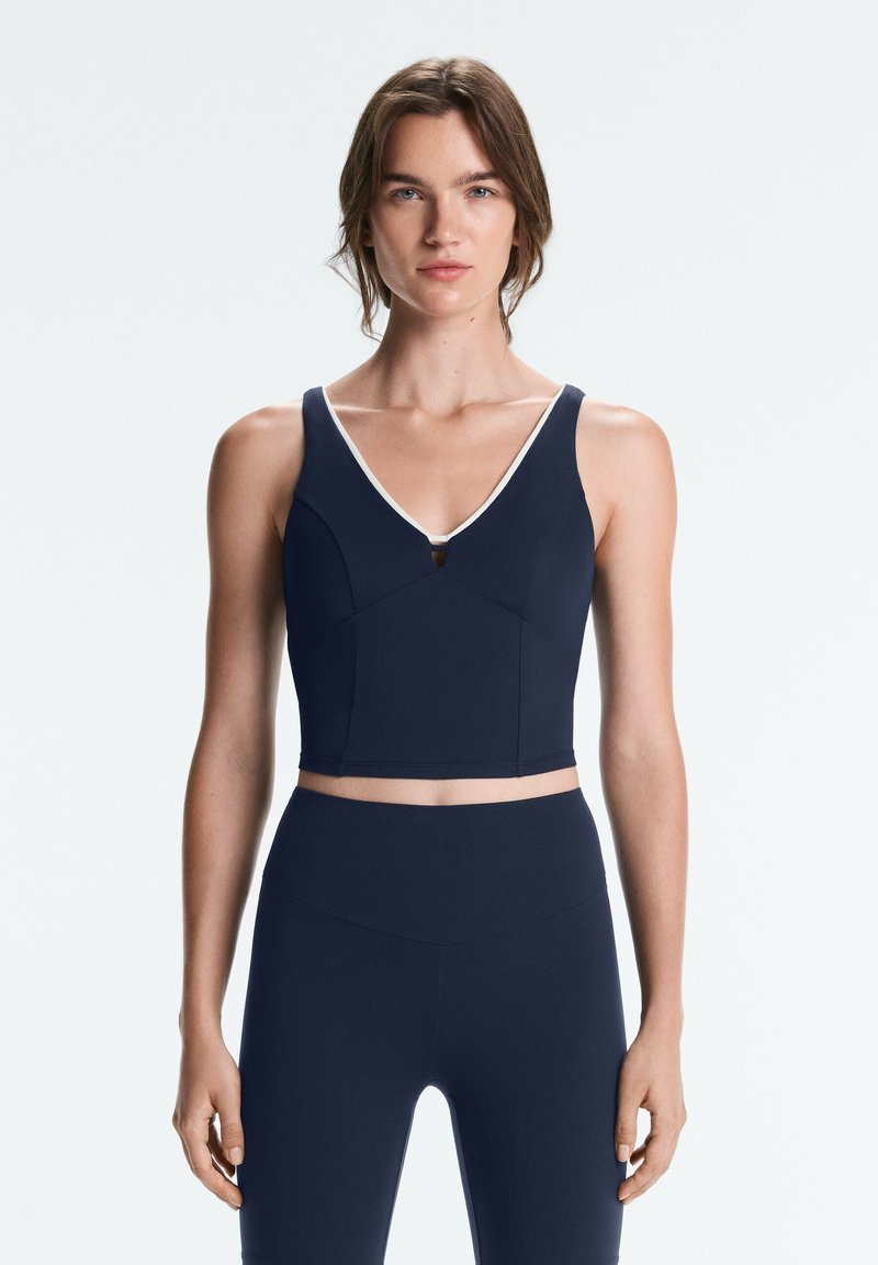 Woman wearing a navy blue sleeveless crop top with white trim and matching high-waisted leggings, standing facing forward on a plain background.