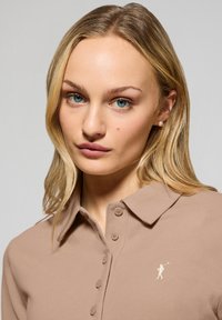 Beige polo tshirt med en strukturerad yta, med krage, tre knappar och en liten broderad logotyp på bröstet.