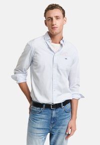 Hellblau-weiß gestreiftes Hemd mit Knöpfen und Kragen, über einem weißen T-Shirt getragen, kombiniert mit blauen Jeans und einem schwarzen Gürtel.