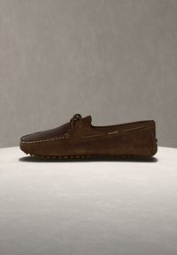 Καφετί μοκασίνι από δέρμα με λευκή ραφή και σόλα από καουτσούκ με κουκκίδες, τοποθετημένο σε φωτεινή επιφάνεια πάνω σε ουδέτερο φόντο.