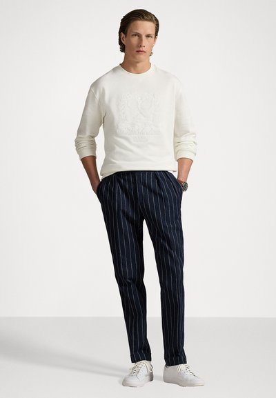 Hanorac alb cu logo-ul de tenis embosat și textul "Wimbledon 2025", asortat cu pantaloni navy cu dungi și sneakers albi.