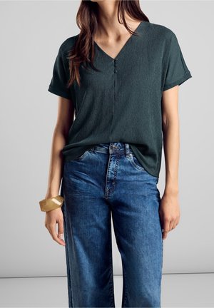 Femme portant un haut vert foncé texturé à manches courtes en V avec trois boutons, associé à un jean bleu taille haute et un bracelet beige.
