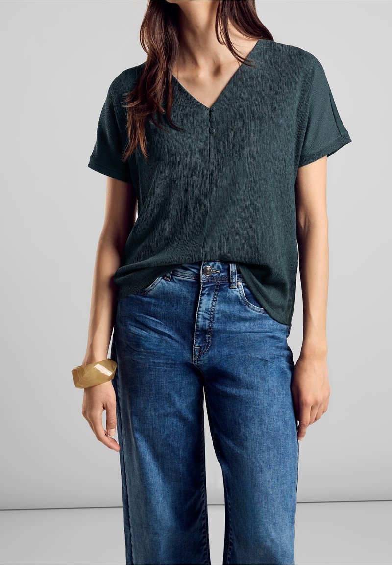 Femme portant un haut vert foncé texturé à manches courtes en V avec trois boutons, associé à un jean bleu taille haute et un bracelet beige.