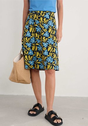 Woman wearing a knee-length skirt with blue and yellow floral pattern, black buckle sandals, holding a woven beige tote bag.
