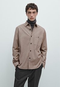 Brun skjorte med knapper lavet af struktureret stof, prydet med en brystlomme og sorte knapper, lagdelt over en sort turtleneck.