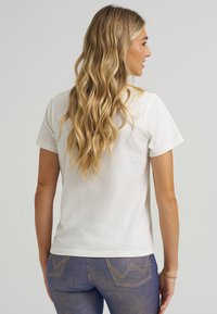 Balta kokvilnas t-kreklus ar apaļu kakla izgriezumu un īsām piedurknēm, skatīts no aizmugures. Mīksta tekstūra, regulāra piegriezuma, apvienots ar zilām džinsu biksēm.