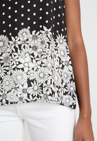 Černý top bez rukávů s bílými puntíky a květinovým potiskem na lemu, kde jsou sedmikrásky a složité linie. Hladká textura.