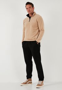 Beige fliisist pullover musta tõmblukuga, kombineeritud mustade dressipükstega ja pruunide tossudega, millel on valged detailid. Sile tekstuur, vabalt voolav lõige.