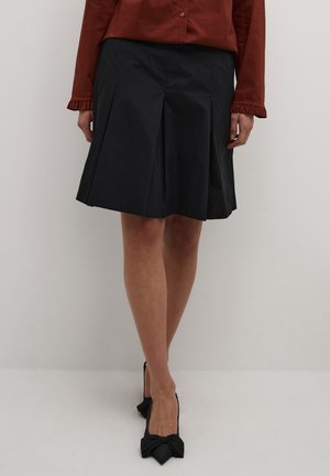 Jupe plissée noire en tissu lisse, longueur genoux, avec une taille cintrée. Portée avec des escarpins noirs à talons hauts ornés de noeuds.