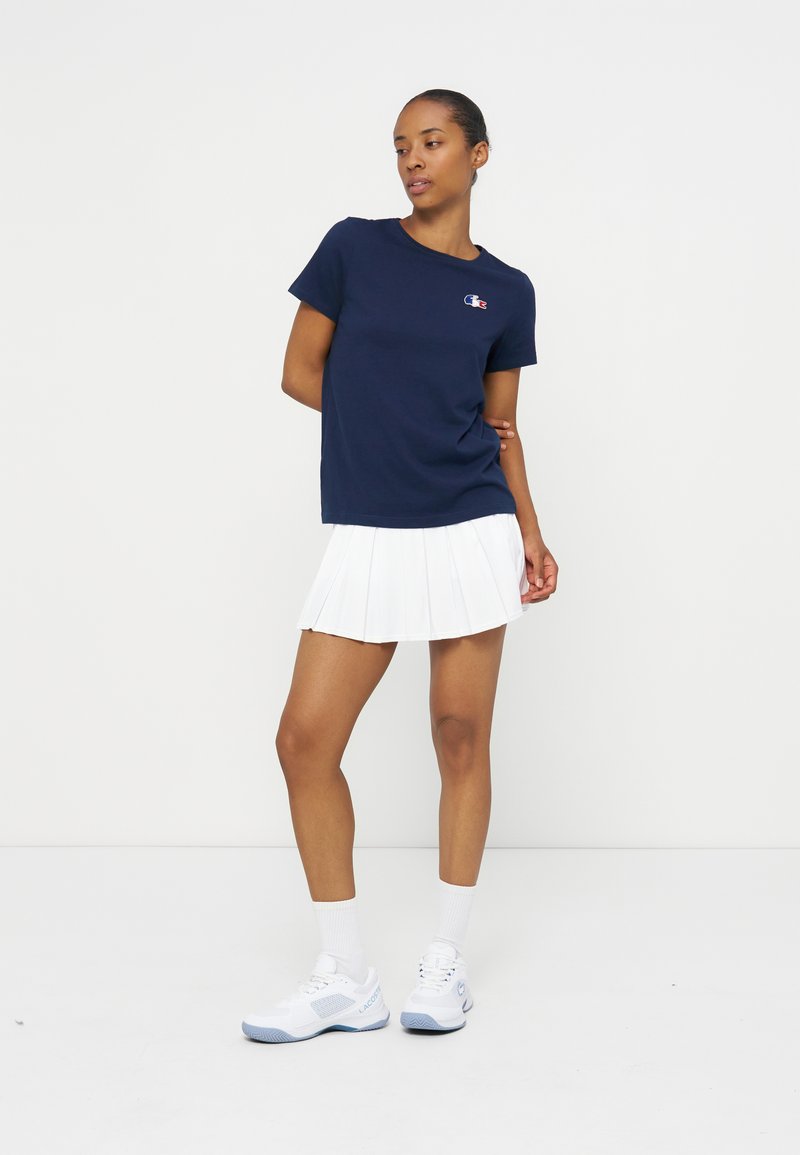 Femme debout, les bras derrière le dos, portant un t-shirt bleu marine, une jupe plissée blanche, des chaussettes blanches et des chaussures de sport blanches.