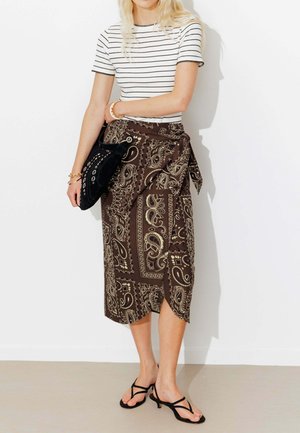 Femme portant un haut rayé à manches courtes, une jupe portefeuille marron à motif paisley, des sandales noires à lanières et tenant un sac à main noir.