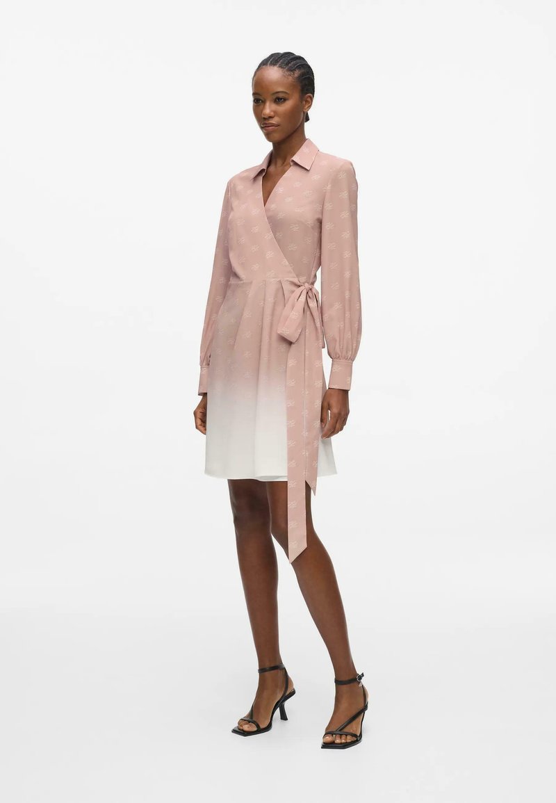 Femme portant une robe portefeuille rose à manches longues avec une jupe ombrée blanche et des sandales à talons hauts noires, debout devant un fond blanc.