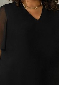 Svart topp med V-ringning, med korta genomskinliga ärmar. Guld kedja som detaljer. Slät textur, enkel siluett.