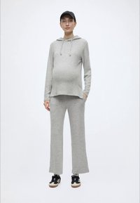 Gaiši pelēks, brīva piegriezuma grūtnieču džemperis ar kapuci un auklu, kopā ar tam atbilstošām platu kāju biksēm. Modele valkā melnas sporta apavus un cepuri.