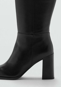 Bota de cuero negro hasta la rodilla con un tacón de bloque texturizado. Diseño elegante, acabado suave, punta redondeada y costuras sutiles.