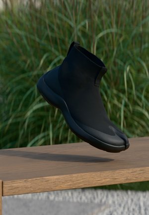 Baskets à chaussettes noires à bout fendu flottant au-dessus d'un banc en bois avec un fond d'herbes hautes, mettant en avant un design minimaliste et élégant.