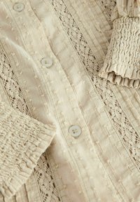 Blouse beige avec un tissu texturé, des motifs en dentelle et des boutons de fermeture. Les manches présentent des détails froncés et volants pour une texture supplémentaire.