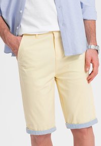 Shorts di cotone gialli con risvolti, caratterizzati da una consistenza liscia e tasche frontali. Abbinati a una camicia azzurro chiaro con bottoni.