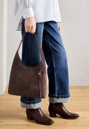 Person wearing cuffed blue jeans and brown ankle boots, holding a dark brown suede handbag with tassel detail.