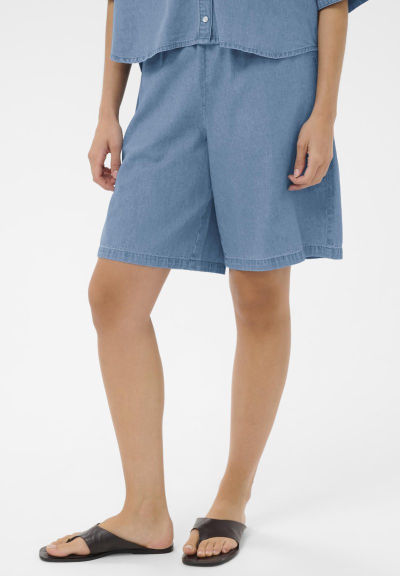 Personne portant un short en denim bleu ample, une chemise boutonnée assortie et des sandales noires à enfiler, debout devant un fond blanc.