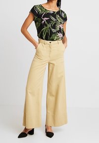 Top negro con flores y mangas cortas; pantalones beige de tiro alto y pata ancha con detalles de costura; tacones negros de punta afilada.