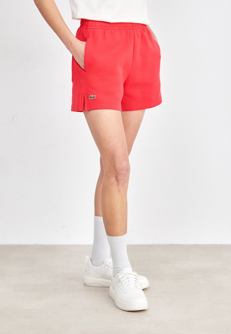 Pantaloni corti rossi con cintura elasticizzata e tasche laterali, caratterizzati da un piccolo logo ricamato. Indossati con scarpe da ginnastica bianche e calzini.