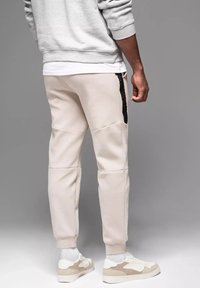 Svetle béžové jogger kalhoty s černým bočním pruhem, s tapered střihem, texturovanou tkaninou a viditelnými švy, nošené s teniskami.