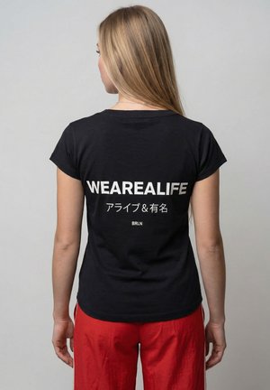 Frau mit langen blonden Haaren, die ein schwarzes T-Shirt mit der Aufschrift "WEAREALIFE" und japanischem Text trägt, kombiniert mit roten Hosen, die von der Kamera weg schaut.