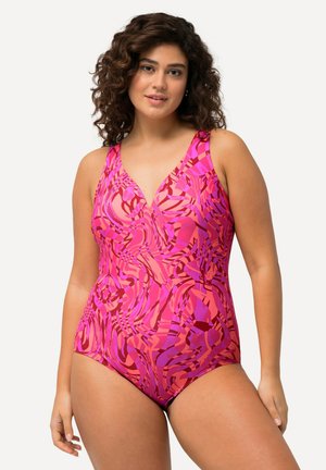 Kvinde med krøllet hår iført en lyserød og rød mønstret badedragt, stående mod en ensartet lys baggrund.