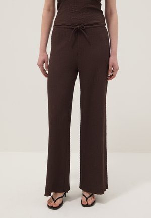 Woman wearing dark brown textured wide-leg pants with a drawstring waist and black heeled thong sandals, standing on a white floor.