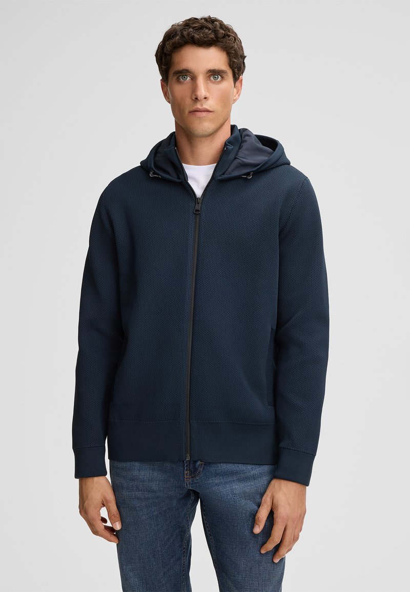 Felpa navy con zip e superficie texturizzata, cappuccio regolabile e tasche laterali. Il tessuto sembra leggero e casual, adatto per essere indossato a strati.