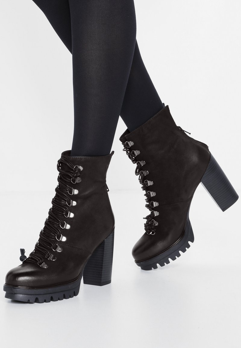 Femme portant des bottines noires à talons épais lacées et des collants opaques noirs, debout devant un fond neutre.