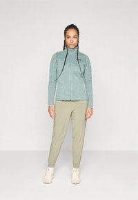 Casaco de lã polar azul claro com gola alta, fecho de correr e mangas compridas, combinado com calças verdes oliva acinturadas e sapatos claros.