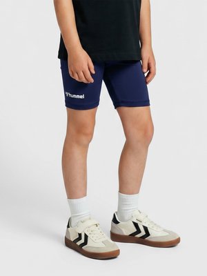 Barn iført marineblå Hummel-shorts, hvide sokker og cremefarvede sneakers med sorte chevroner på en lysegrå baggrund.